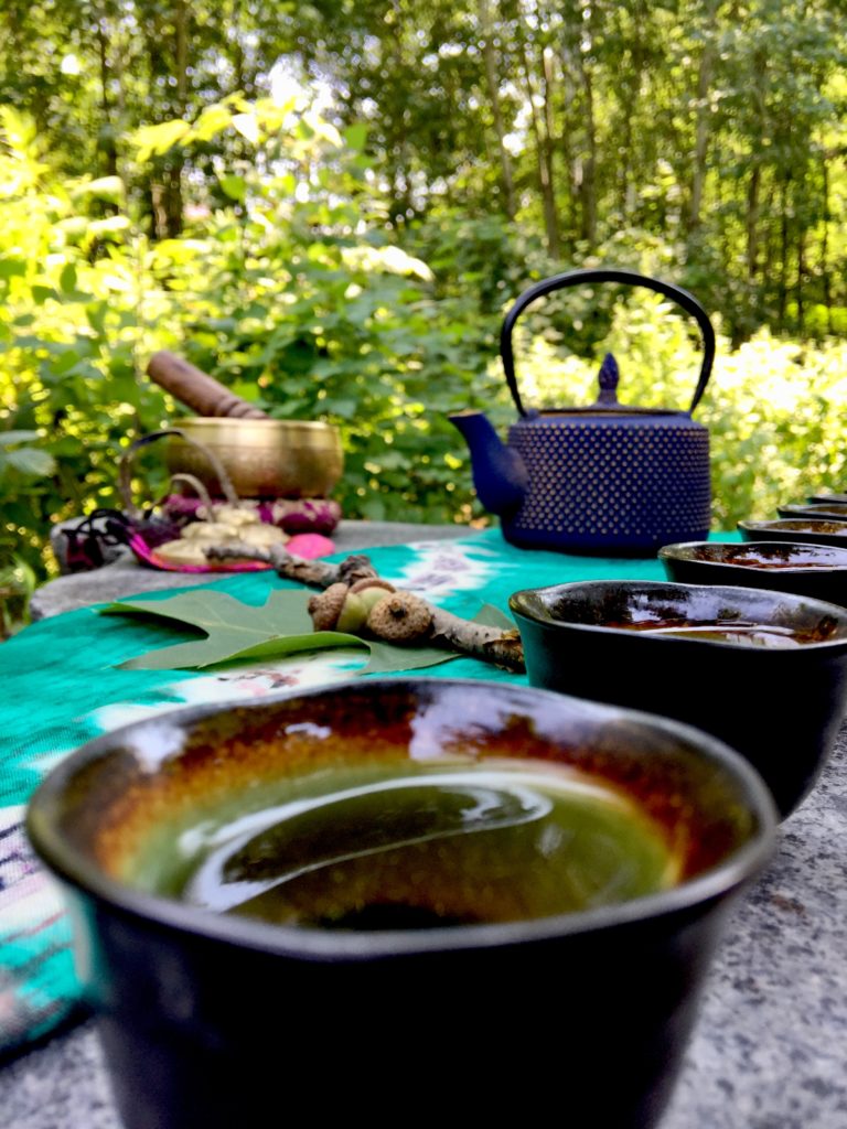A small teacup filled with tea in the foreground, several more teacups and a teapot in the background. The setup is outside, and lush greenery surrounds the scene.