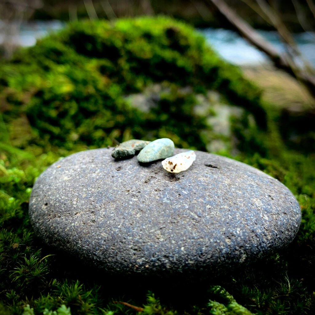 A smooth, grey stone, topped with three very small stones in a row: grey, light green, and white. The stones rest upon a bed of moss, next to a river.