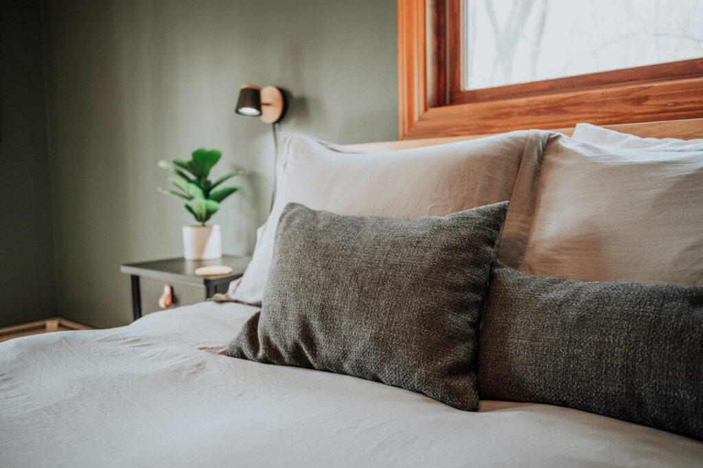 Photo of a bed with cozy bed pillows in two shades of grey, with a green plant on a side table in the background.